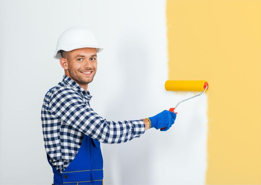 A decorator is painting a wall yellow with a roller and smiling. He is wearing a hard hat and blue overalls.