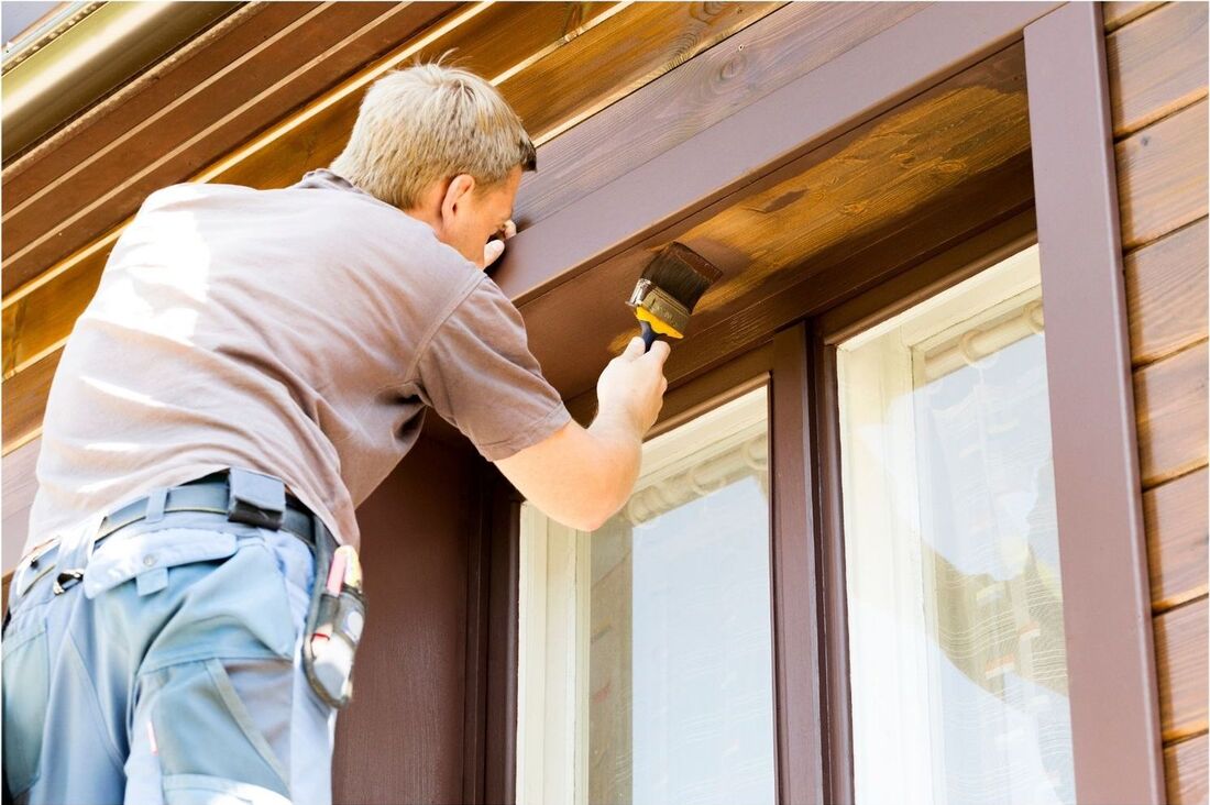 A painter is painting the outside of a wooden window frame dark brown.