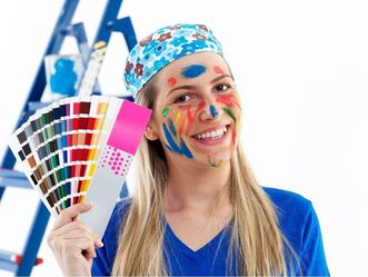 A lady is smiling and holding up some colour sample cards. She is wearing a blue top and a patterned bandana and her face is covered in different colours of paint.
