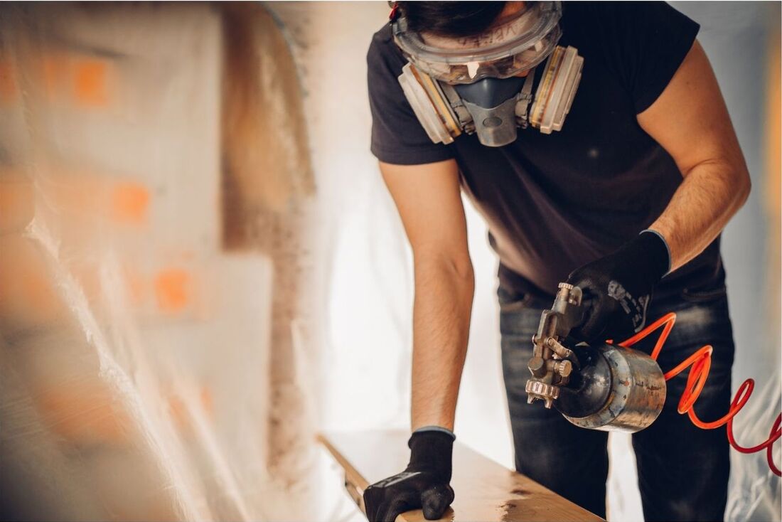 A decorator is spray painting a wooden panel. He is wearing a protective mask and black gloves, as well as a black t-shirt.